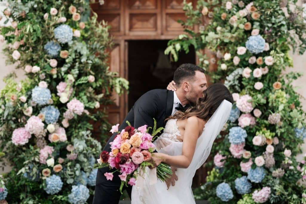 Fotografia di una coppia di sposi che si bacia, circondata da un arco floreale di rose e ortensie, durante il matrimonio decorato e organizzato da ChiaraB Events Wedding Planner.