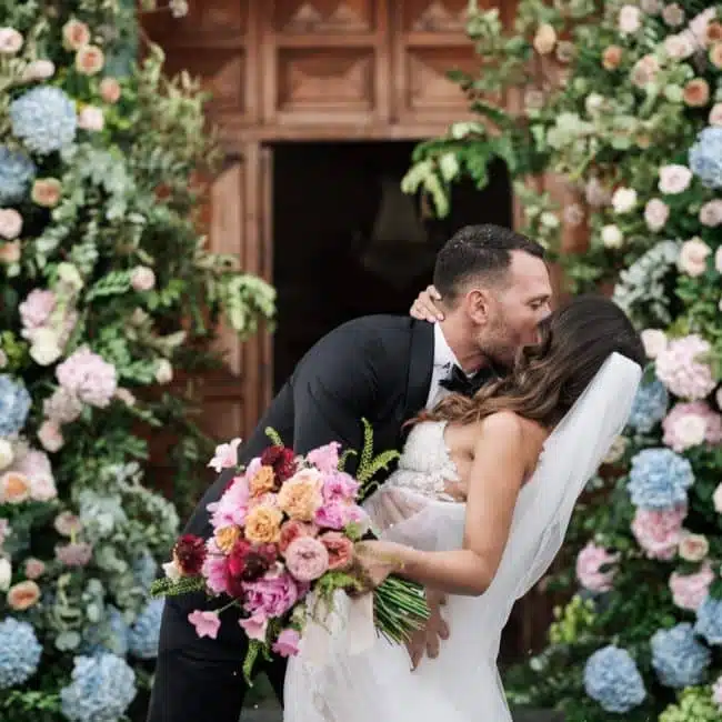 Fotografi av ett gift par som kysser varandra, omgivna av en blomsterbåge av rosor och hortensior, under ett bröllop dekorerat och organiserat av ChiaraB Events Wedding Planner.