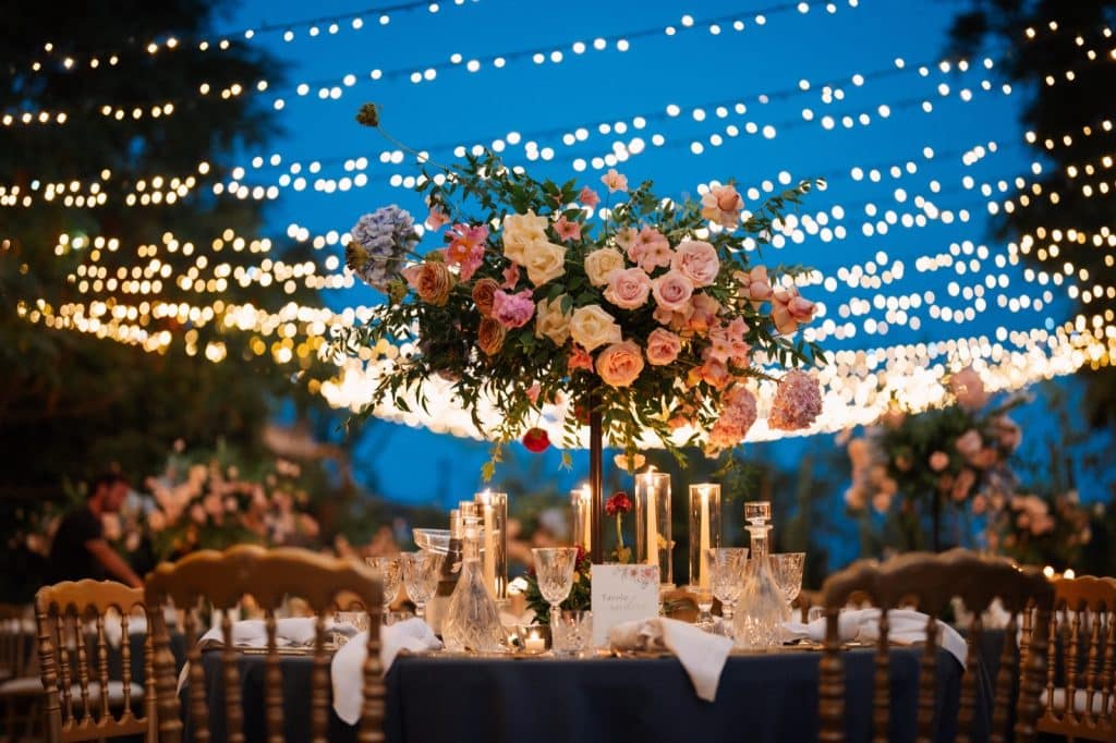Elegante decorazione nuziale con centro tavola floreale di rose e amaryllis, illuminata da candele, sotto stringhe di luci (fairy lights) per matrimoni indimenticabili, servizio wedding planner ChiaraB Events.