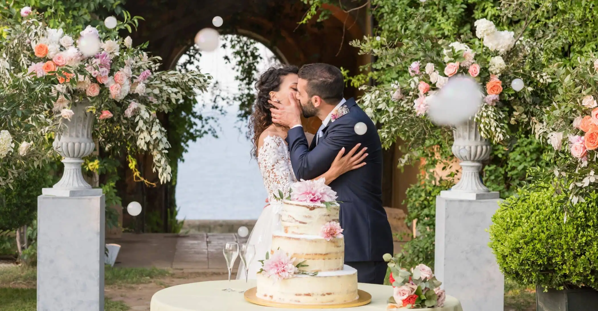 Photograph of a married couple kissing during their outdoor wedding, with elegant floral decorations and a wedding cake, perfect for a wedding planner service.