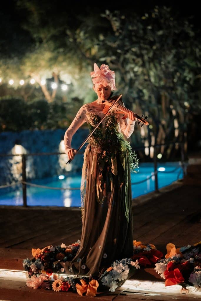 Elegante artista che suona il violino in un matrimonio all'aperto circondata da fiori colorati, sotto luci soffuse, con un'atmosfera romantica e raffinata, perfetta per un servizio di wedding planning esclusivo.
