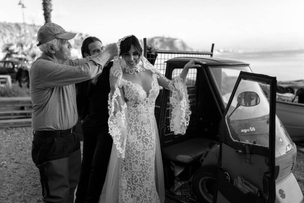Elegante sposa in abito da matrimonio che si prepara con assistenza di professionisti per il giorno del matrimonio, in una location all'aperto vicino al mare, promuovendo servizi di wedding planner.