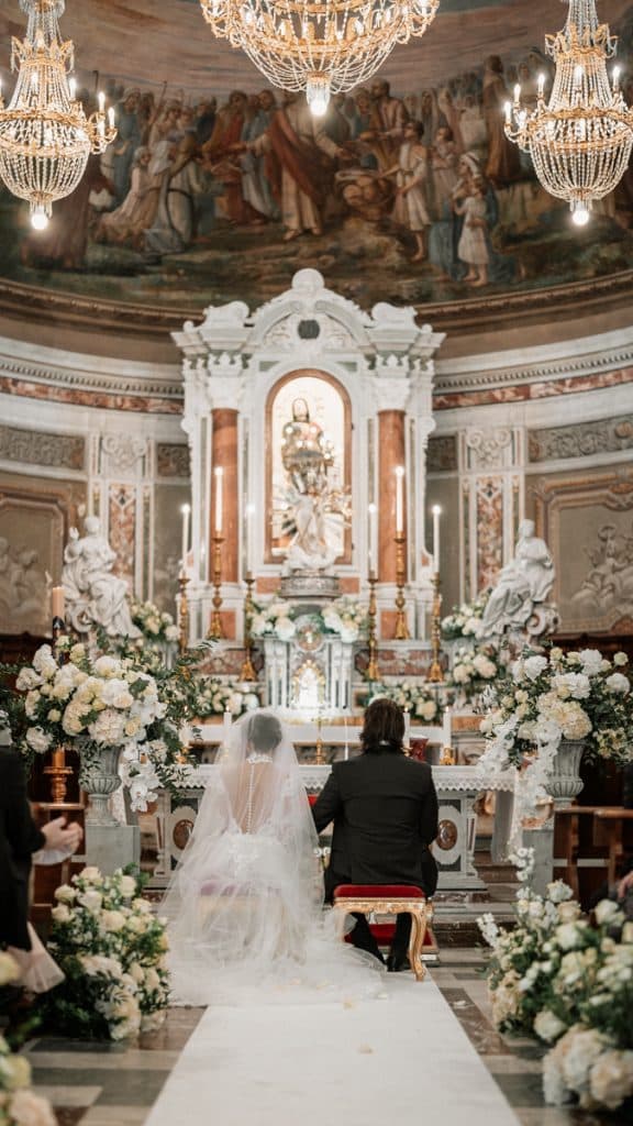 Fotografia di una cerimonia di nozze in chiesa, con decorazioni floreali bianche, altare barocco e due sposi seduti durante il rito, illuminato da lampadari di cristallo, perfetta per pianificare matrimoni eleganti e cerimoni sacre.