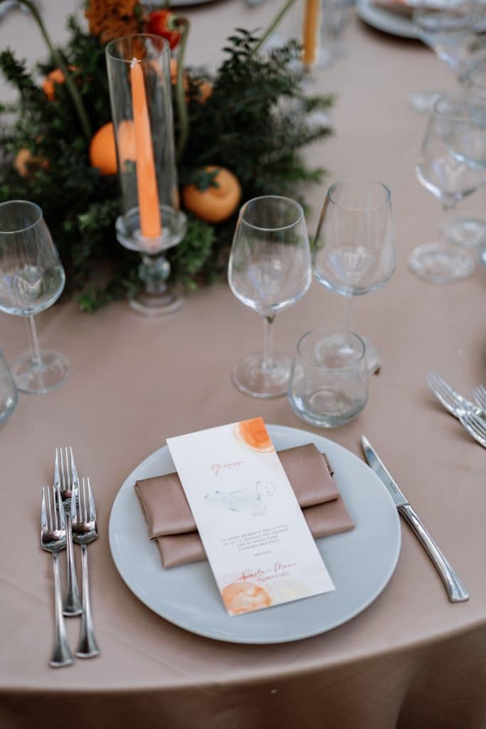 Elegante tavolo da matrimonio con tovaglia beige, sottopiatti, calici di cristallo, posate e menu personalizzato, decorato con fiori e candele, progettato da ChiaraB Events Wedding Planner, ideali per eventi speciali e matrimoni di lusso.