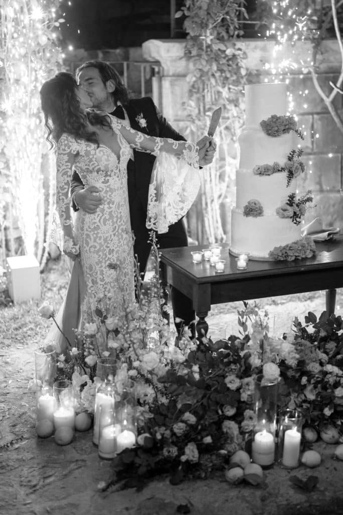 Ciotola di una torta nuziale decorata con fiori e foglie, illuminata da candele, con una coppia felice che si bacia e si abbraccia durante un matrimonio elegante e romantico, pianificato da ChiaraB Events Wedding Planner.