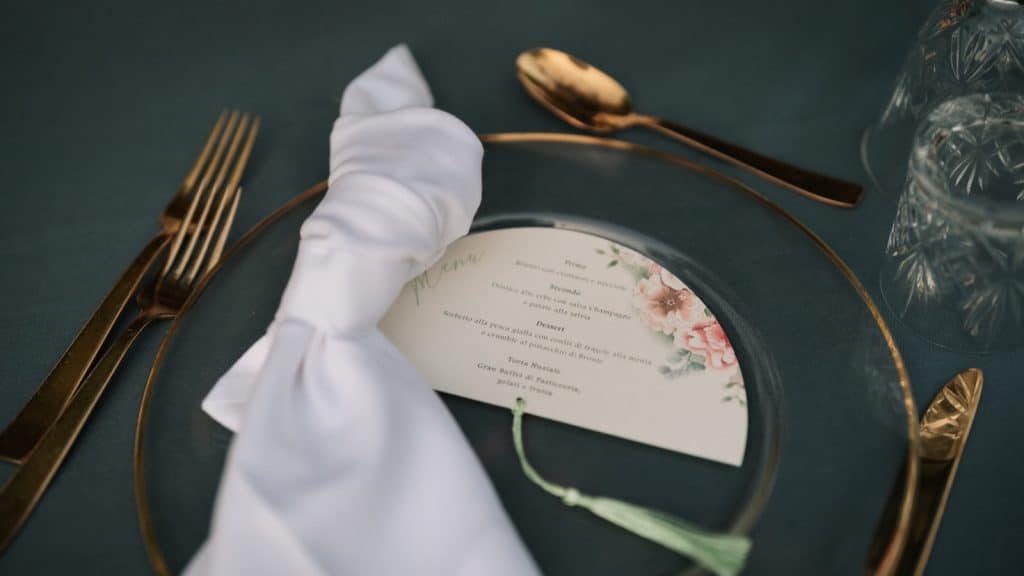 Menu di nozze elegante su tavola decorata con posate oro, tovaglia blu scuro e bicchieri cristallo, perfetto per wedding planner e organizzatore di matrimoni.