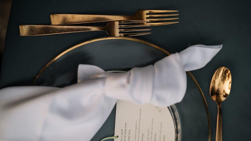 Eleganza tavola nuziale con posate in oro, tovagliolo bianco e menu di matrimonio su piatto scuro, perfetta per wedding planner e allestimenti matrimoni raffinati.
