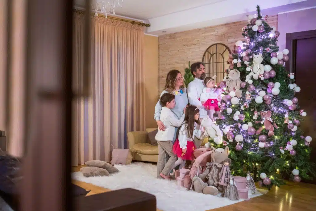 Albero di Natale decorato con ornamenti rosa e bianco, circondato da regali e statuette, con famiglia felice che lo ammira, in un'accogliente sala addobbata per le festività natalizie.