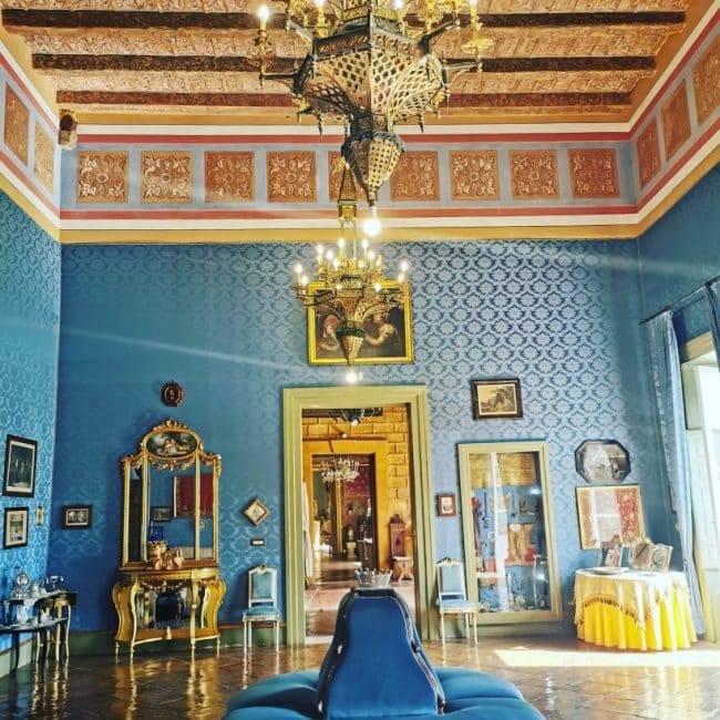 Elegante salone di matrimonio con pareti blu, soffitto decorato con travi in legno e affreschi, lampadari in oro e cristallo, arredamento di lusso e opere d'arte, ideale per eventi di nozze raffinati.