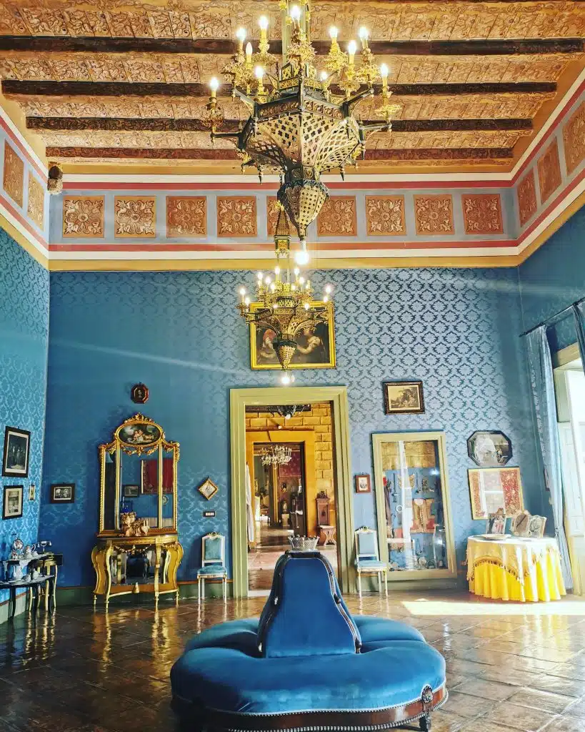 Um elegante salão de casamento com paredes azuis, um teto decorado com vigas de madeira e frescos, lustres de ouro e cristal, mobiliário de luxo e obras de arte, ideal para eventos de casamento sofisticados.