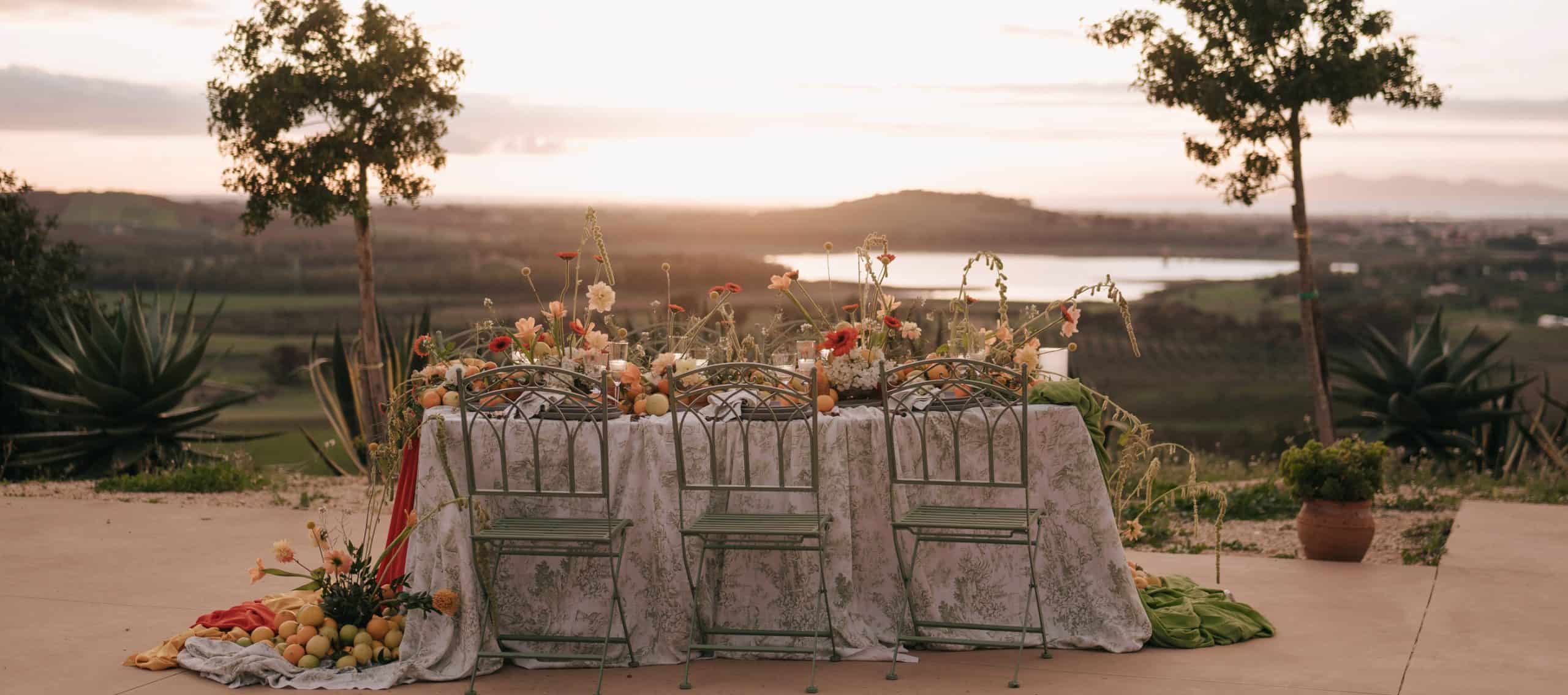 Tavolo per matrimonio all'aperto decorato con fiori e frutta, con vista panoramica al tramonto, perfetto per eventi romantici e sofisticati.