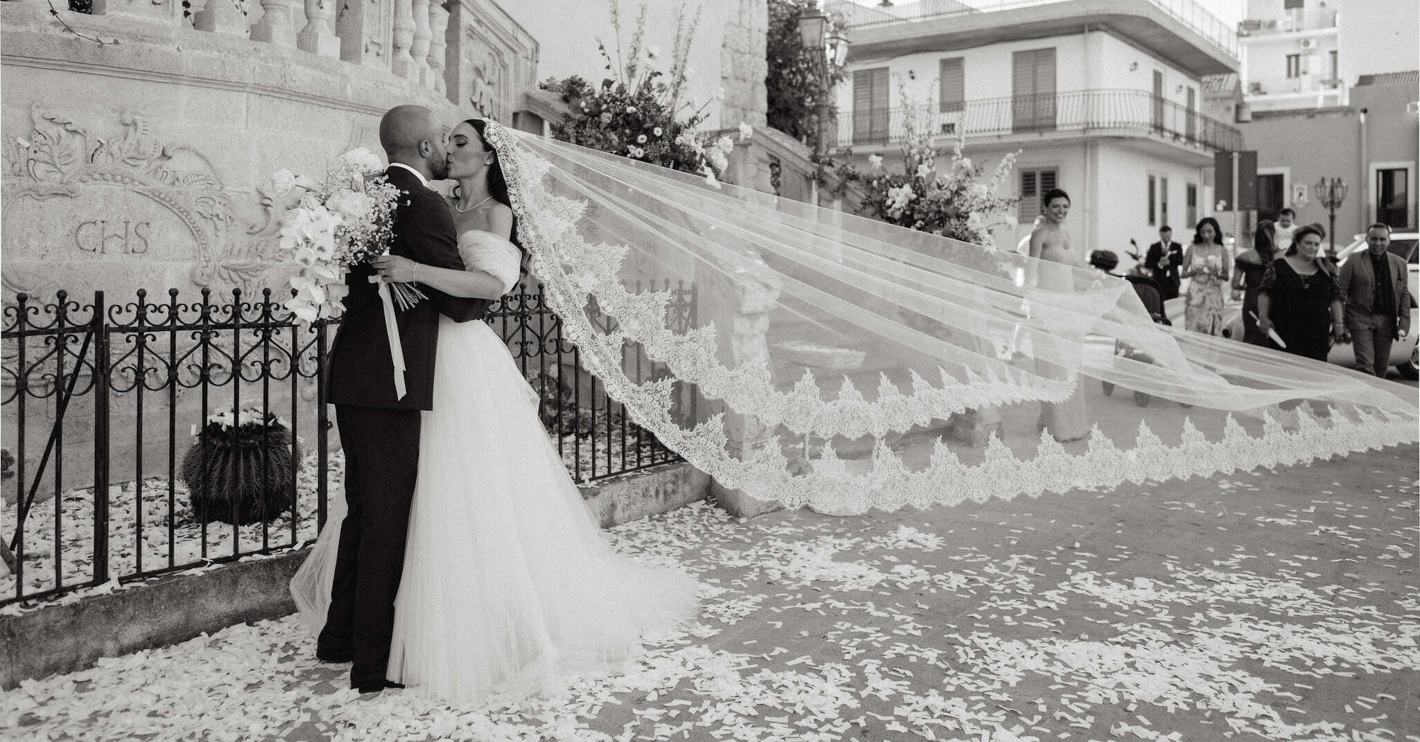 Alberto e Valeria si scambiano un bacio romantico durante il loro matrimonio, con un lungo velo bianco e un abito da sposa elegante, in un contesto urbano e festoso.