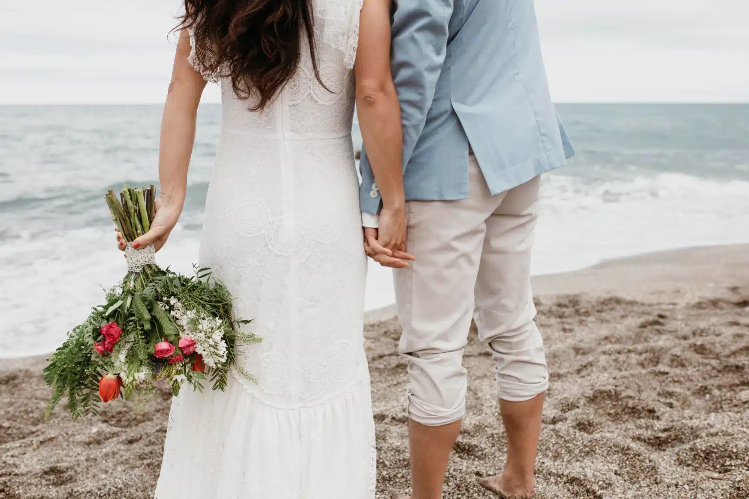 Pris, alt-tekst: Nygifte på stranden med blomsterbuket, romantisk udendørs bryllupsbillede, bryllupsarrangør Chiara B Events.