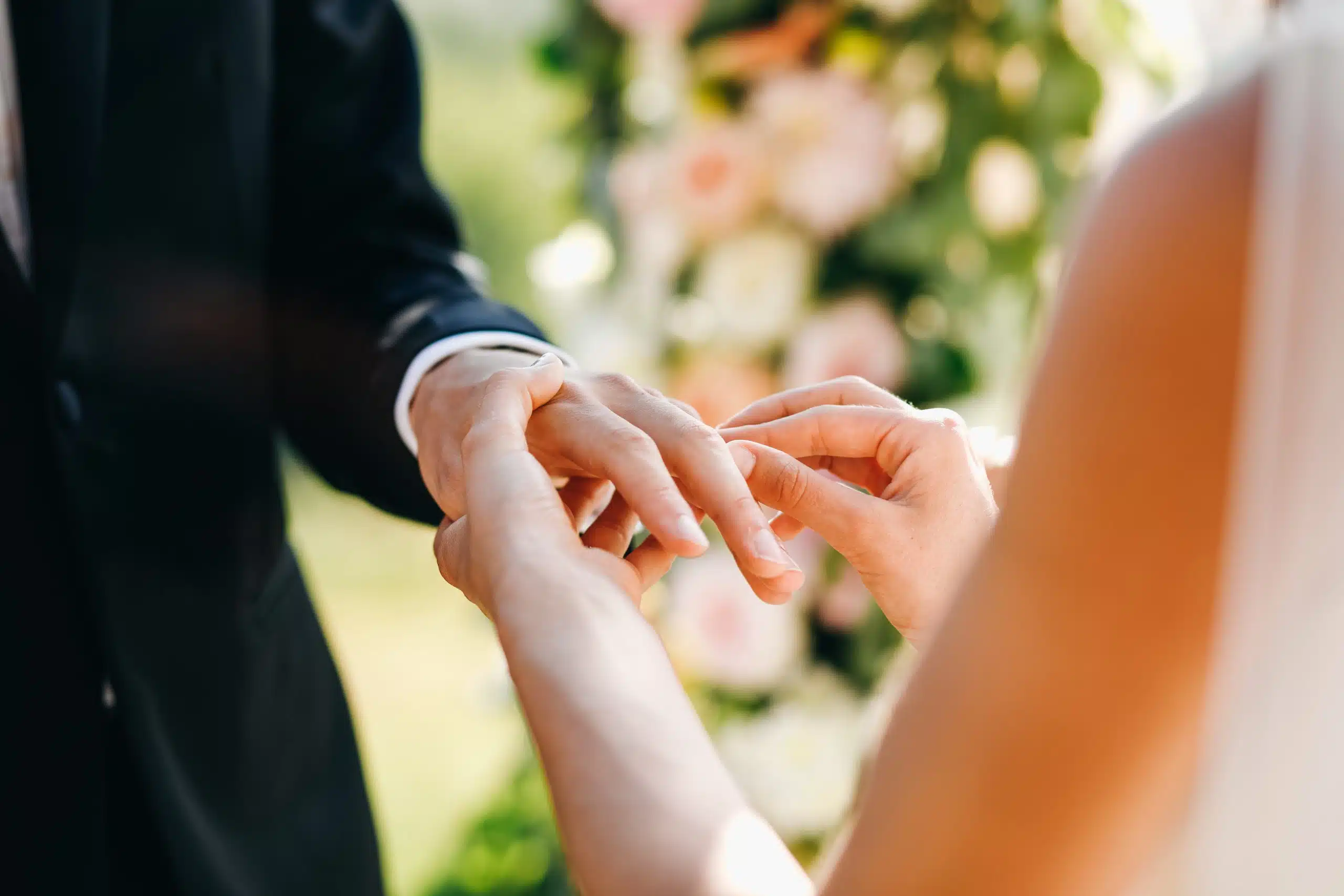 Anello di nozze, coppia che si scambia gli anelli durante un matrimonio, servizio wedding planner, evento romantico, rilassato e elegante, decorazioni floreali in sfondo.