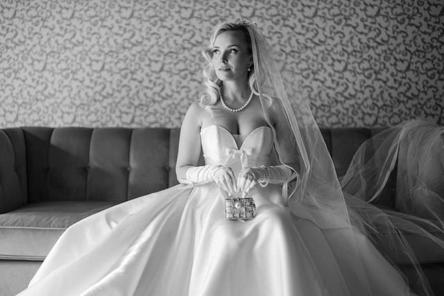 Sposa in abito da sposa bianco, seduta su un divano, con dettagli raffinati e un velo, in un ambiente elegante.