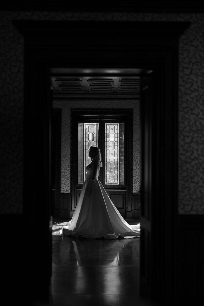 Elegante sposa in abito da matrimonio che si staglia davanti a una grande finestra con vetrate decorative, in un ambiente classico e raffinato, perfetto per servizi fotografici di wedding planning.