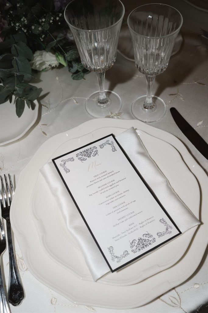 Menù di nozze elegante con dettagli decorativi raffinati, posizionato su un piatto bianco con bicchieri di cristallo e decorazioni floreali, perfetto per matrimoni sofisticati e personalizzati.