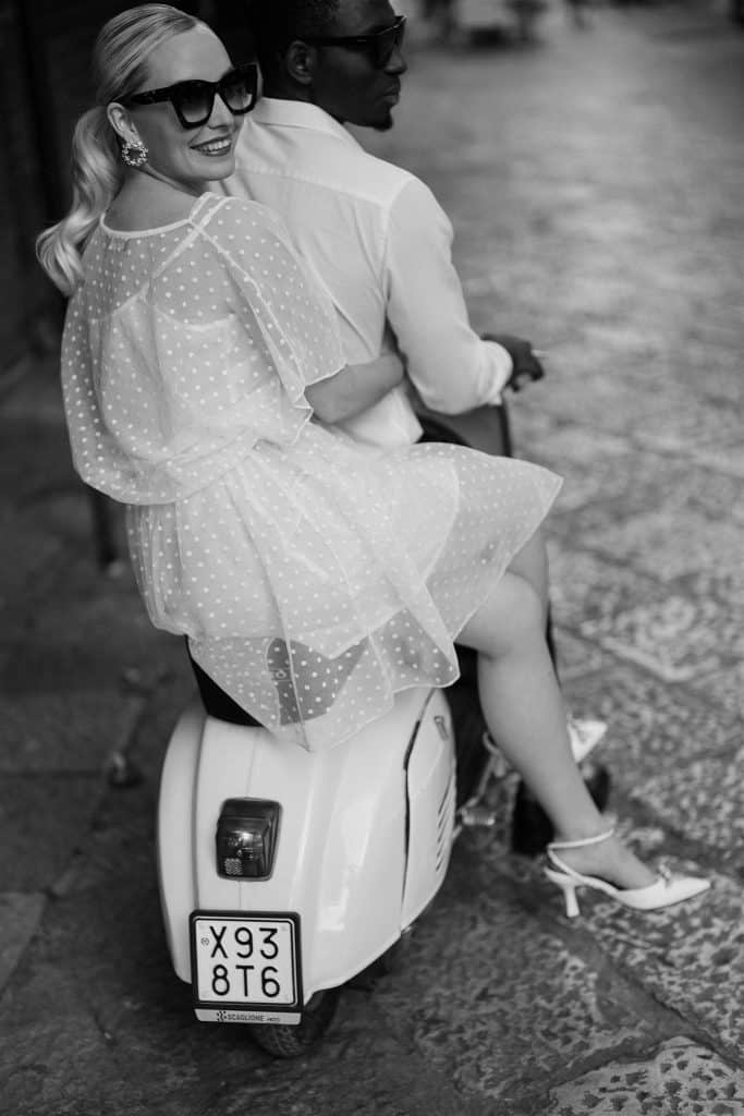 Una coppia felice su uno scooter, donna con abito leggero e occhiali da sole, uomo con camicia bianca, in un ambiente urbano. Perfetto per servizi di wedding planning e matrimoni eleganti.
