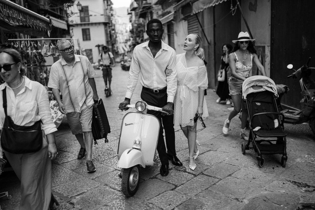 Una coppia di sposi in abiti eleganti cammina per le strade di una città storica, circondata da passanti e negozi, con un'atmosfera romantica e vivace.