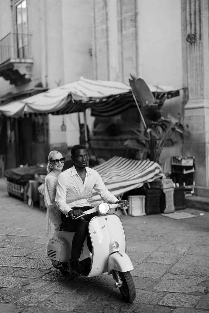 Una coppia su uno scooter in una strada storica con bancarelle e edifici antichi, in un'immagine in bianco e nero che trasmette un'atmosfera romantica e vintage.