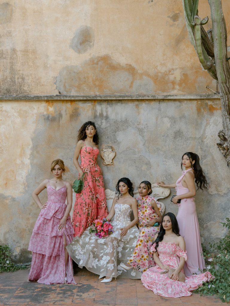 Elegant group of women in floral and pastel evening dresses, posing in front of an ancient wall, perfect for wedding planning services and special events in a romantic and sophisticated style.