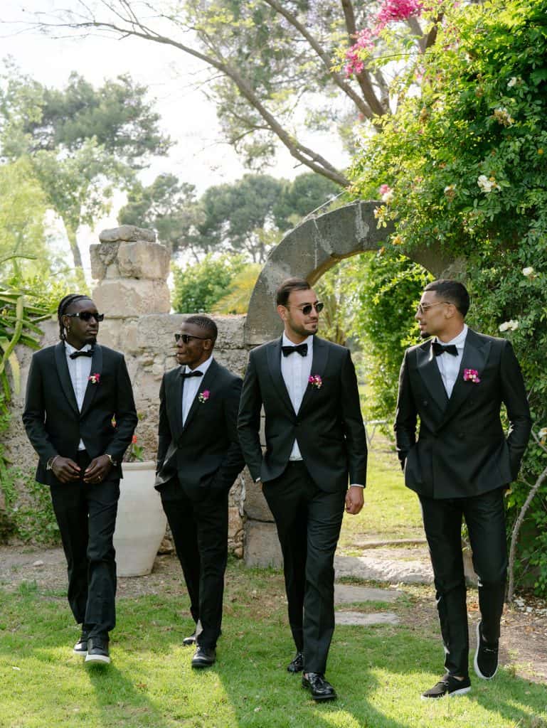 Elegant men in black formal suits with bow ties, walking in a lush garden, perfect for a wedding or special event, under an arch of flowers and plants.