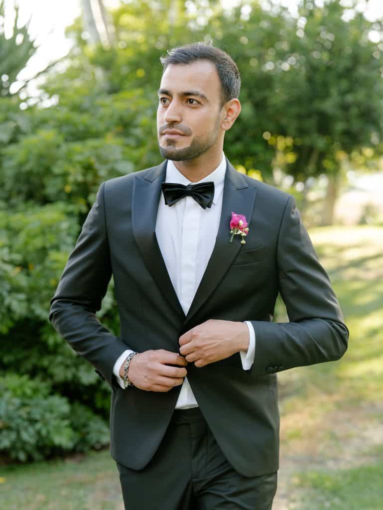 Elegant groom in a black suit with bow tie, ready for the wedding, in a green garden with soft lights, representing a professional wedding planning service by ChiaraB Events.