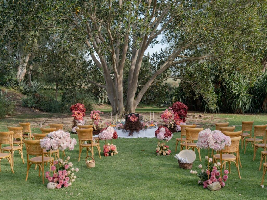 An outdoor area set up for a wedding with wooden chairs, pink and white floral decorations, under a large tree, perfect for romantic ceremonies and special events.