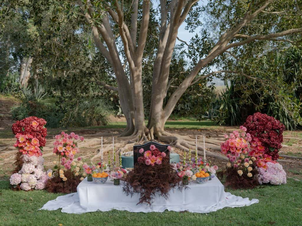 Elegant wedding setup with a table decorated with colorful flowers under a large tree in a natural garden, perfect for romantic events and outdoor ceremonies.