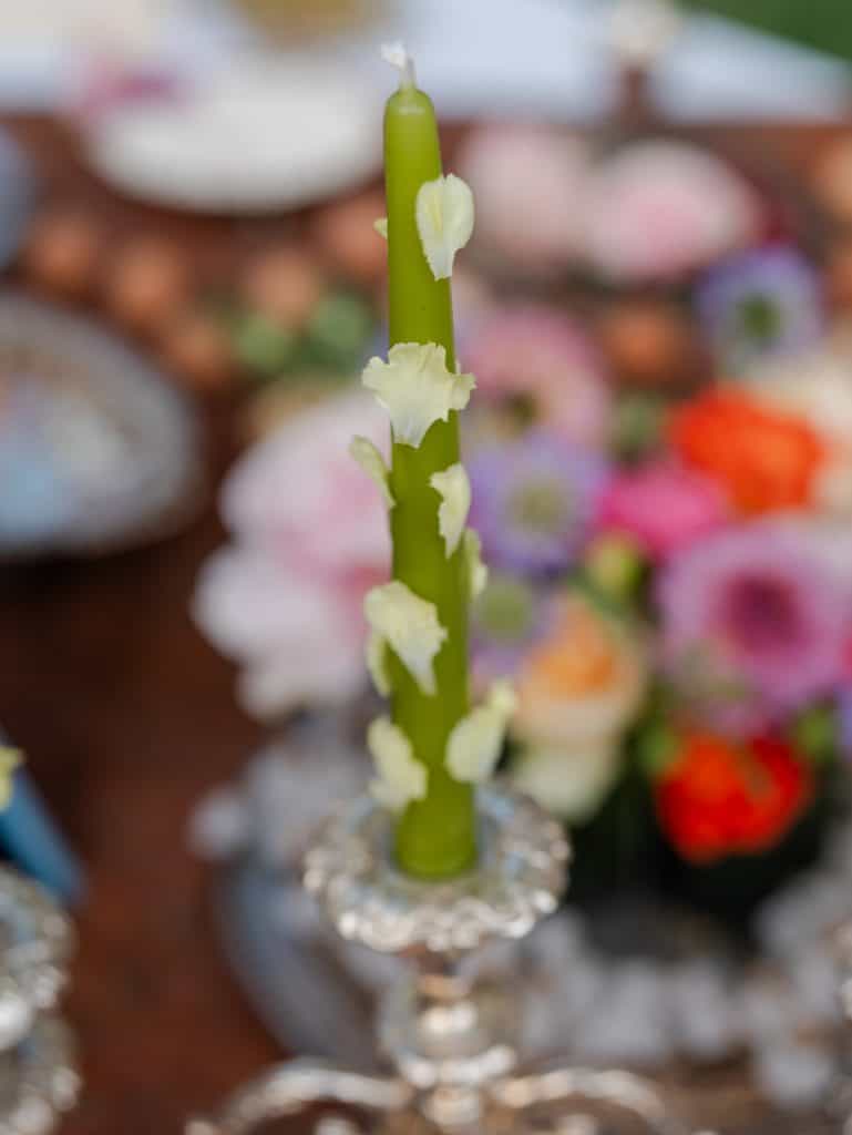 A green candle decorated with white flowers, perfect for weddings and elegant events, on a table decorated with colorful flowers and refined details.