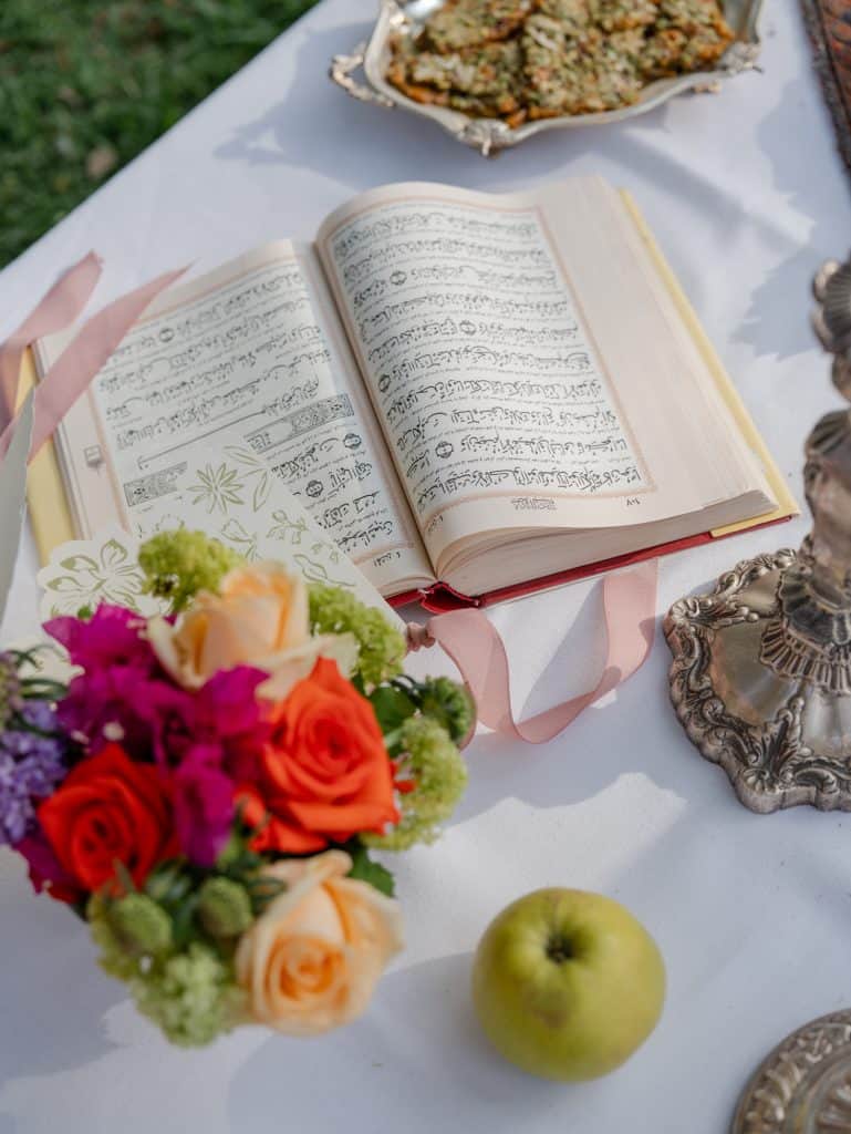 Open prayer book with colorful flowers, green apple, and decorative objects on a wedding table, wedding planning service by ChiaraB Events.