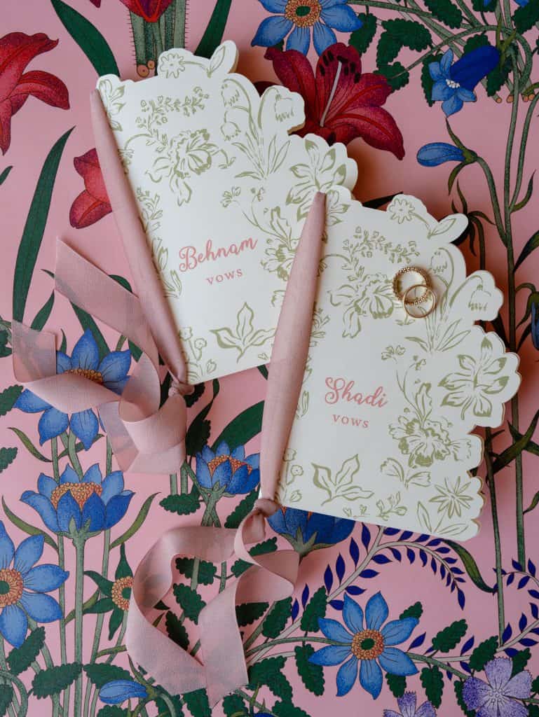 Elegant personalized wedding invitations with golden floral details, accompanied by wedding rings and pink silk ribbons, on a background of colorful flowers, perfect for a romantic and refined wedding.