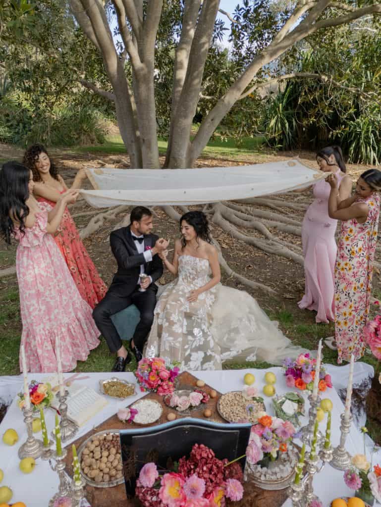 An outdoor wedding with bride and groom and bridesmaids under a large tree, decorated with flowers and candles, in a natural and romantic setting.