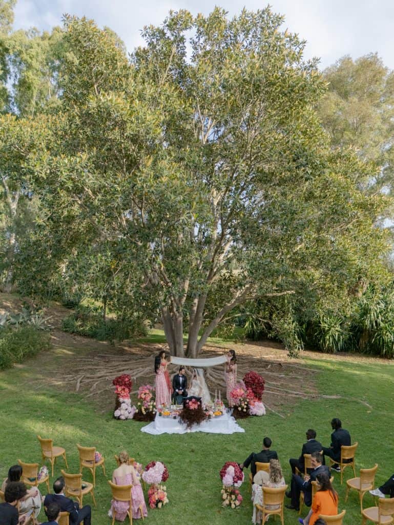 Outdoor ceremony under a large tree with floral decorations and seated audience, wedding event organized by ChiaraB Events.
