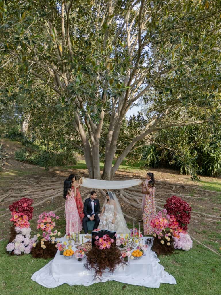 An ALT tag: Outdoor wedding ceremony under a large tree with colorful floral decorations and seated bride and groom, planned by ChiaraB Events Wedding Planner, in a natural and romantic environment.