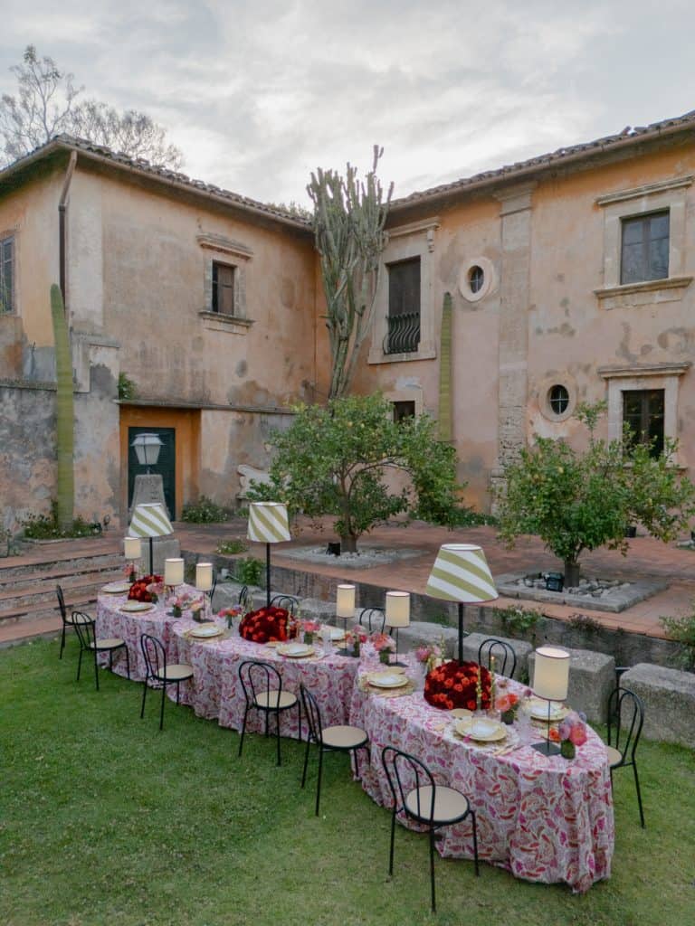 Outdoor table decorated for a wedding, with red flowers and candles, in a historic courtyard with stone buildings and green plants, perfect for elegant and romantic events.