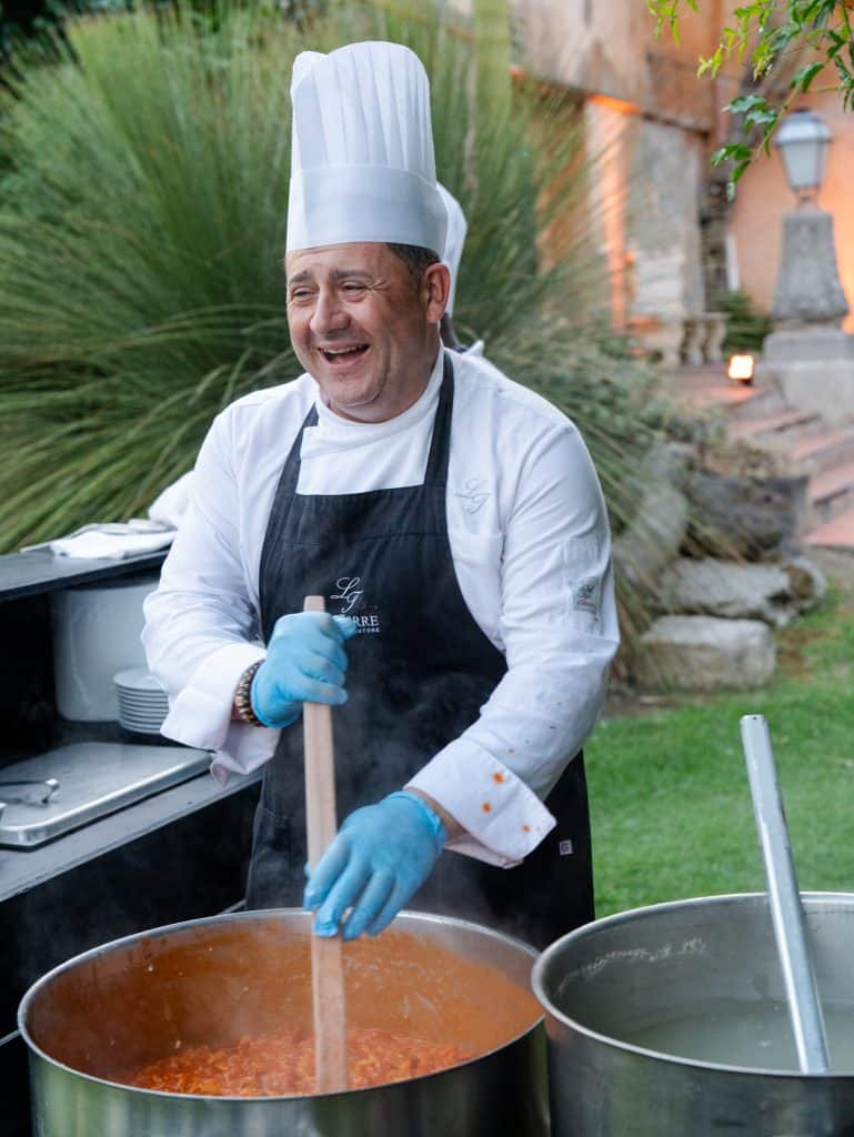 A smiling chef in professional attire cooking a hot soup in an outdoor setting, with plants and stones in the background, representing the excellence of ChiaraB Events as a wedding planner and c.
