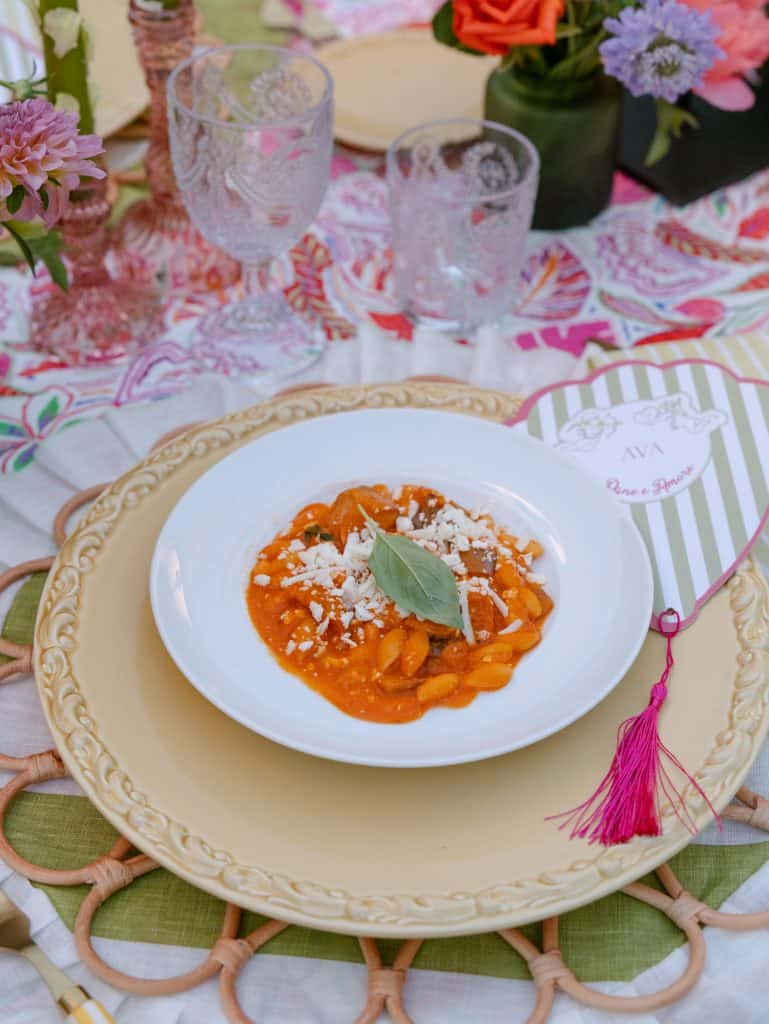 Plate of pasta with tomato sauce, decorated with a basil leaf, on an elegant white plate, part of a wedding table with colorful flowers and crystal glasses, perfect for events.