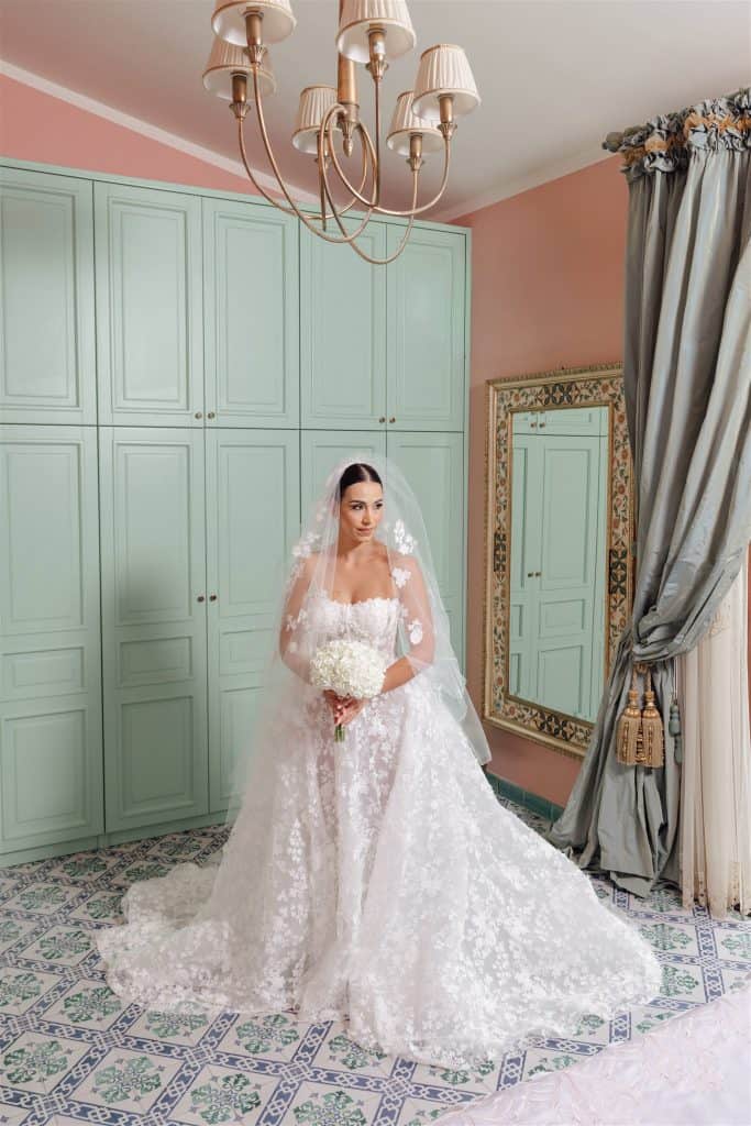 Sposa in abito da matrimonio con velo e bouquet di fiori, in una stanza elegante con pareti rosa, armadio verde pastello, grande specchio e tende grigie, pronta per il matrimonio.