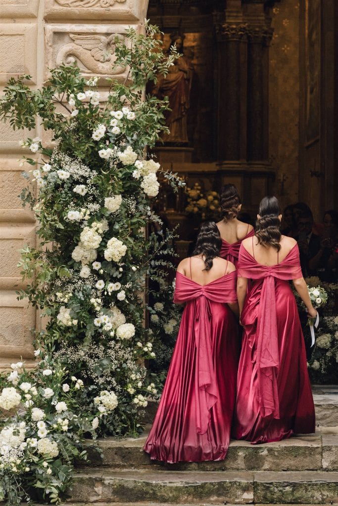 Eleganti damigelle in abiti rosa scuro con dettagli plissettati, che si avvicinano all'ingresso di una chiesa decorata con un arco floreale di rose bianche e verde, pronta per un matrimonio.