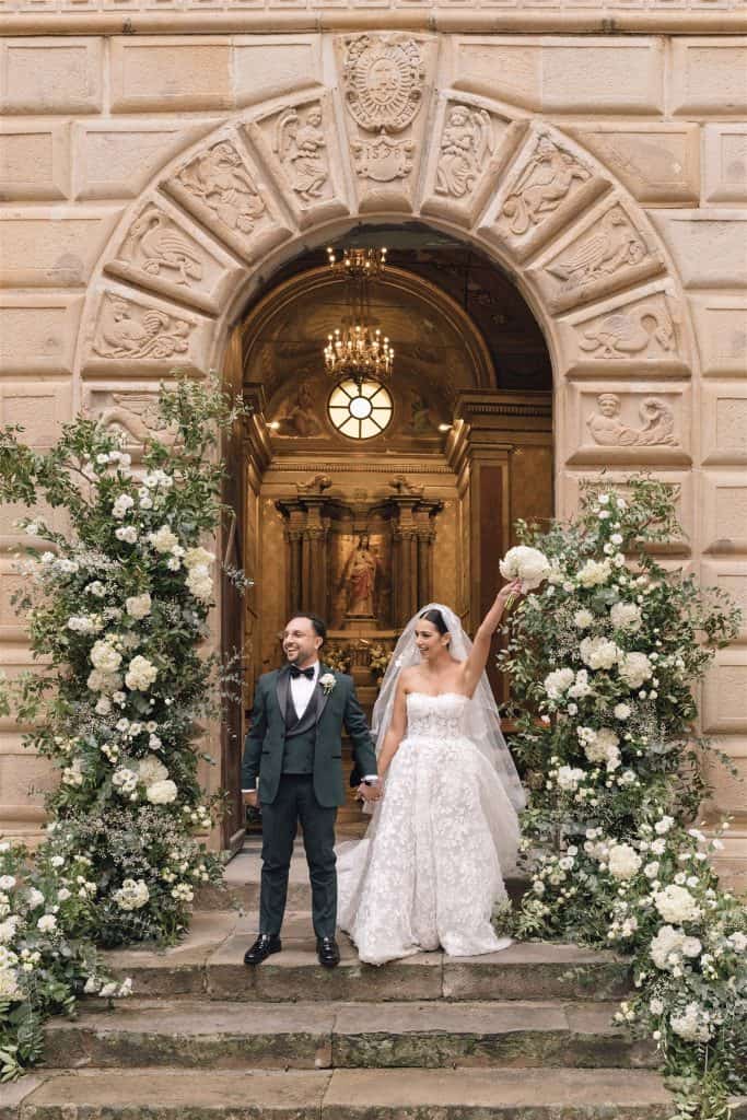 Sposi felici davanti all'ingresso di una chiesa storica decorata con fiori bianchi e verdi, celebrando un matrimonio elegante e romantico, perfetto per servizi di wedding planning e organizzazione eve.