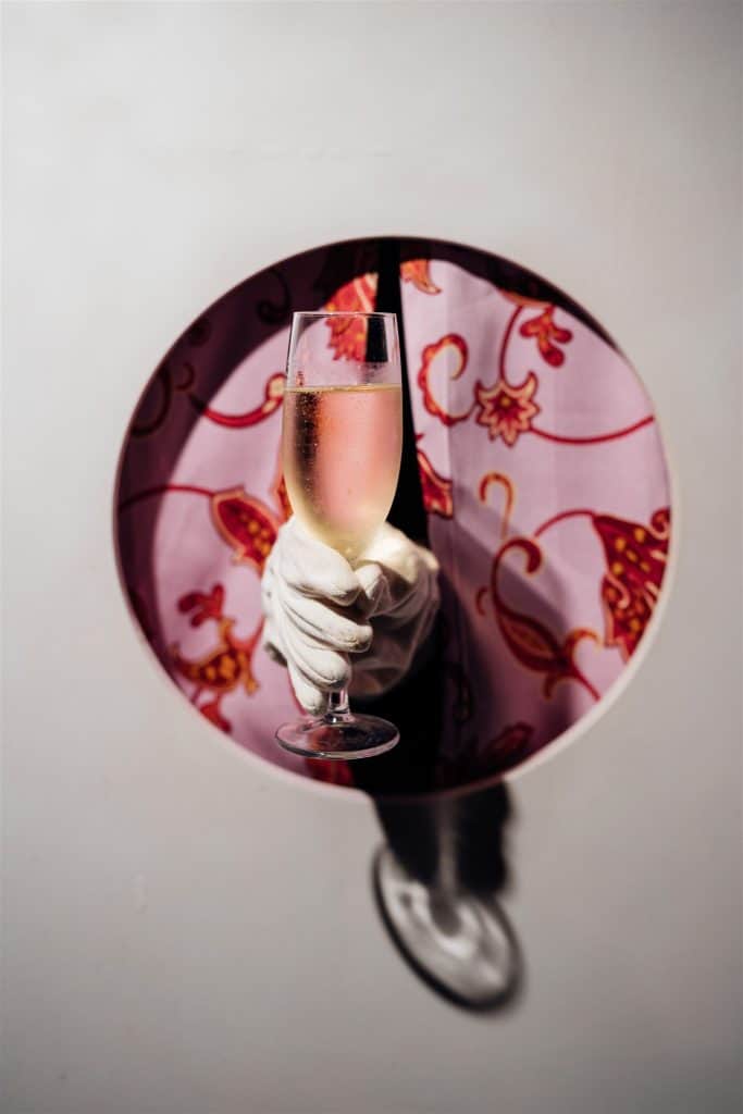Una mano con guanto bianco tiene un calice di champagne rosa, visto attraverso un foro circolare in una parete bianca, con sfondo decorativo floreale rosso e rosa.