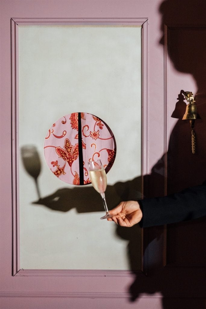Un bicchiere di champagne tenuto da una mano, riflesso in uno specchio decorato con motivi floreali rossi e oro, in un ambiente elegante e raffinato, ideale per eventi e matrimoni.