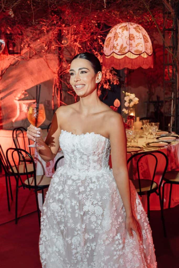 Elegante sposa in abito da matrimonio bianco con dettagli floreali, sorride e tiene un bicchiere di cocktail in un ambiente festoso e raffinato, perfetto per servizi di wedding planning e eventi speci.