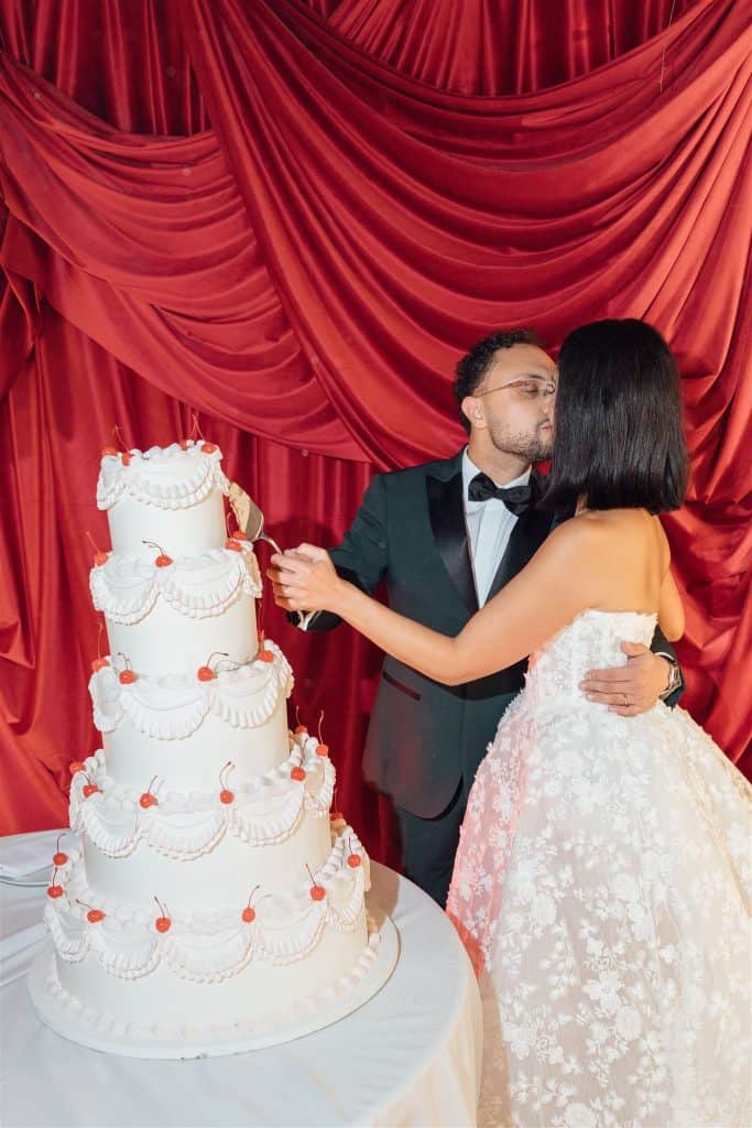 Torta nuziale elegante con decorazioni di panna e ciliegie, coppia di sposi che si scambia un bacio davanti a un drappeggio rosso, perfetta per matrimoni romantici e pianificati da ChiaraB Events.