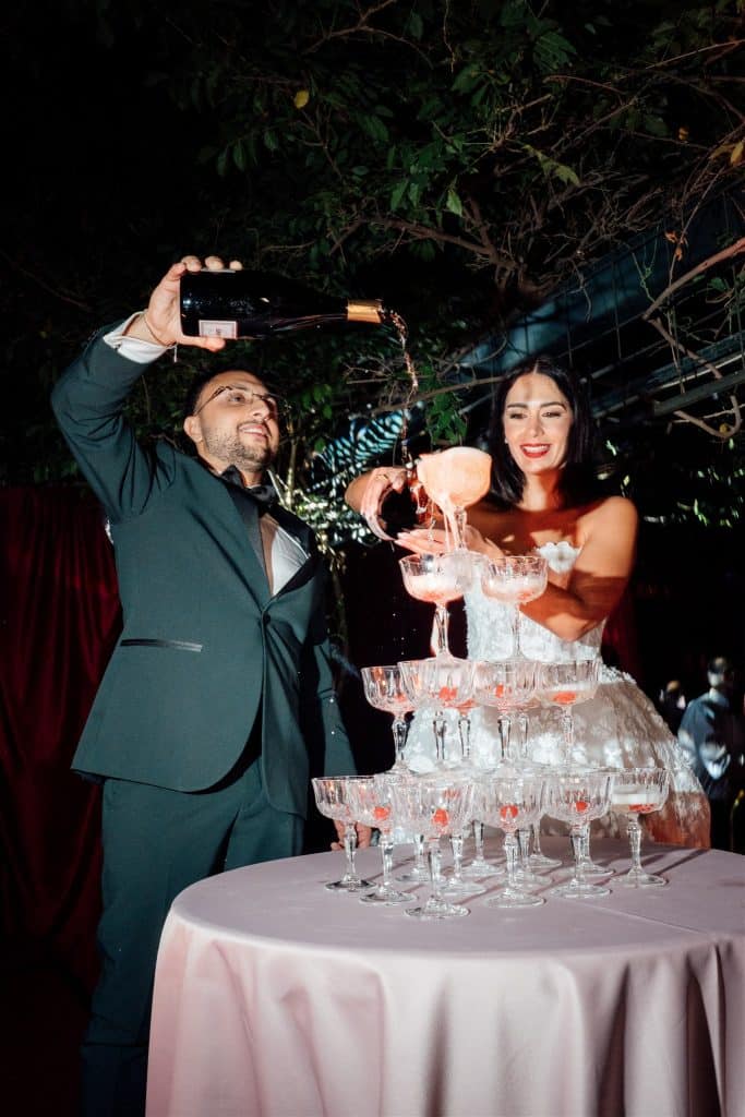 Tappo di champagne che viene versato in una torre di bicchieri da sposa e sposo durante un matrimonio elegante all'aperto.