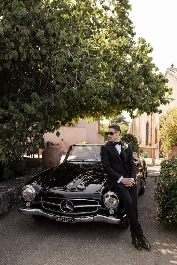 Un uomo elegante in abito da sera con occhiali da sole si appoggia a una Mercedes-Benz classica nera, sotto un grande albero verde, in un ambiente raffinato e tranquillo, perfetto per servizi di weddi.