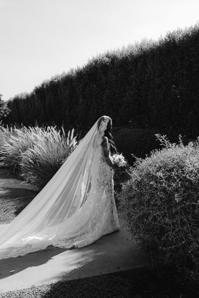 Elegante sposa in abito da matrimonio con velo lungo, in un giardino curato, perfetta per servizi di wedding planning e organizzazione matrimoni in Italia.