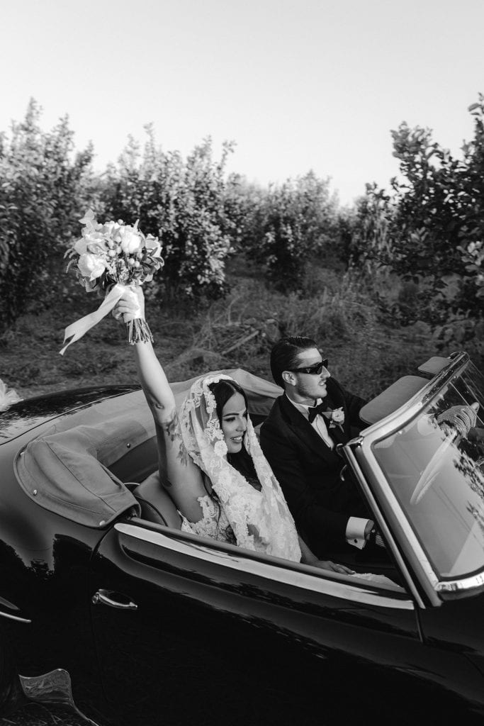 Coppia di sposi in auto d'epoca durante il matrimonio, con la sposa che tiene un bouquet e indossa un velo, in un paesaggio naturale all'aperto, in stile elegante e romantico.