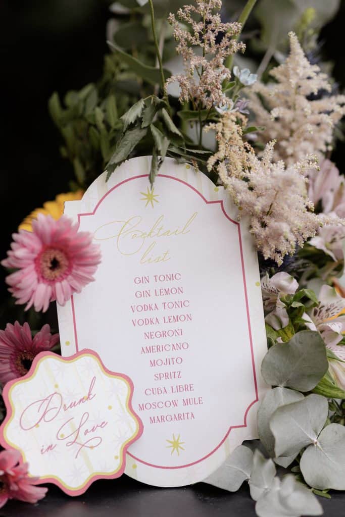 Un bouquet floreale elegante con un cartellino con lista di cocktail, decorato con fiori rosa, bianchi e foglie verdi, ideale per eventi e matrimoni organizzati da ChiaraB Events.